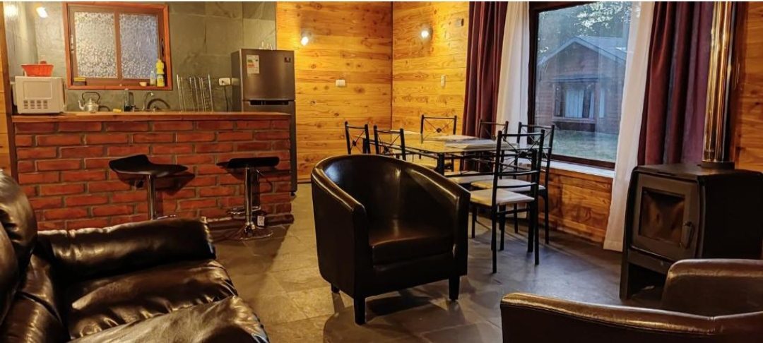 Living de cabaña con sillones, comedor, cocina y estufa a leña